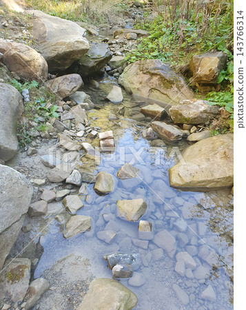 Clear shallow mountain stream flowing between large boulders and scattered stones, with blue sky reflections and green plants on the banks Clear shallow mountain stream flowing between large boulders and scattered stones, with blue sky reflections and green plants on the banks 143766314