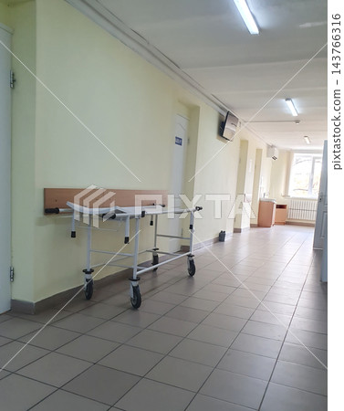 Empty hospital corridor with a wheeled medical stretcher parked by a yellow wall, tiled floor, ceiling lights and a window at the end Empty hospital corridor with a wheeled medical stretcher parked by a yellow wall, tiled floor, ceiling lights and a window at the end 143766316