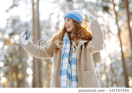 Young woman in a bright hat and scarf with a phone in a snowy forest. Travel, vacation concept. Young woman in a bright hat and scarf with a phone in a snowy forest. Travel, vacation concept. 143767494
