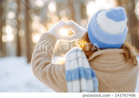 Woman in hat and scarf against backdrop of snowy winter forest holds hands in the shape of a heart. Woman in hat and scarf against backdrop of snowy winter forest holds hands in the shape of a heart. 143767496