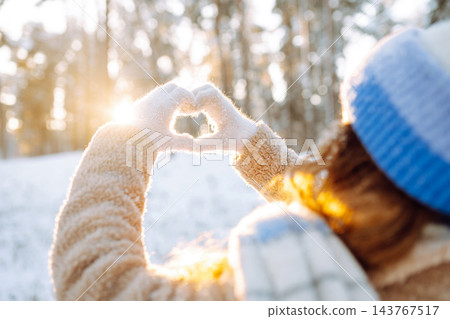 Woman in hat and scarf against backdrop of snowy winter forest holds hands in the shape of a heart. Woman in hat and scarf against backdrop of snowy winter forest holds hands in the shape of a heart. 143767517