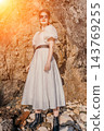 Woman cliff dress standing against a rugged coastal wall in a white vintage style gown under warm sunset light 143769255