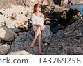 Woman coast rocks sitting on rugged beach shore during bright sunny day wearing white shirt and sunglasses 143769258