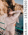 Woman sea cliff young lady in beige knitwear looks out at the blue ocean from a rocky coastline during a sunny vacation 143769260
