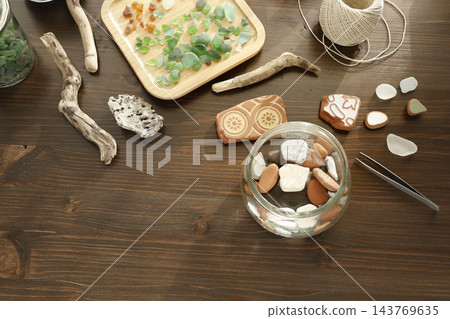 Glass jar with smooth beach pebbles and sea pottery shards on dark wood table. Flatlay coastal treasures with driftwood twine cord tweezers. Sustainable zero waste ocean DIY home decor project. Glass jar with smooth beach pebbles and sea pottery shards on dark wood table. Flatlay coastal treasures with driftwood twine cord tweezers. Sustainable zero waste ocean DIY home decor project. 143769635