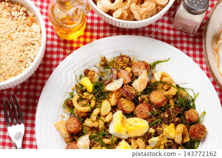 Brazilian feijao tropeiro with sausage, pork rinds and eggs served on a white ceramic plate on a table with a checkered tablecloth. 143772162