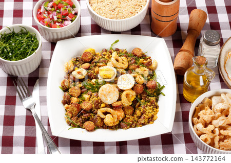 Brazilian feijao tropeiro with sausage, pork rinds and eggs served on a white square plate on a table with a checkered tablecloth. Brazilian feijao tropeiro with sausage, pork rinds and eggs served on a white square plate on a table with a checkered tablecloth. 143772164