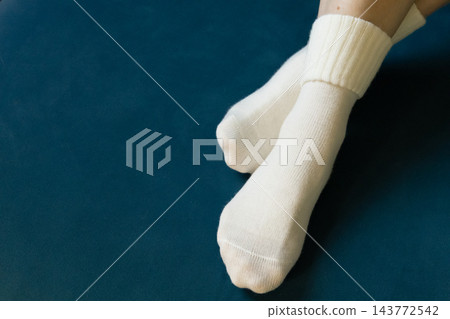 Woman crossing legs in white socks. Girl legs in cozy wool socks on blue background. Cozy autumn lifestyle. Woven socks on woman legs. Woman holding feet on blue sofa. Winter fashion. Woman crossing legs in white socks. Girl legs in cozy wool socks on blue background. Cozy autumn lifestyle. Woven socks on woman legs. Woman holding feet on blue sofa. Winter fashion. 143772542