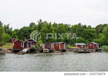 Traditional red wooden boathouses along peaceful Nordic shoreline with rustic fishing piers and lush forest backdrop Traditional red wooden boathouses along peaceful Nordic shoreline with rustic fishing piers and lush forest backdrop 143772674