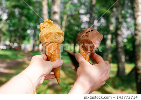 Two hands holding colorful ice cream cones with chocolate and pistachio scoops in sunny summer park 143772714