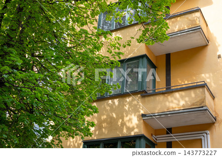 Yellow apartment building facade with green tree branches in sunlight Yellow apartment building facade with green tree branches in sunlight 143773227