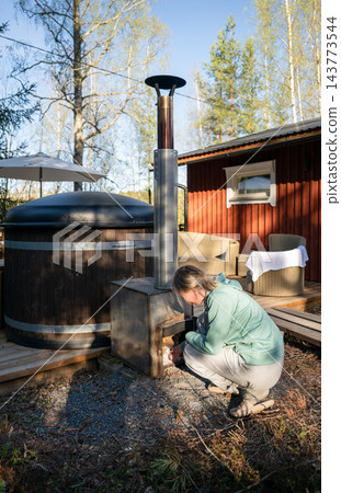 Woman heating traditional outdoor hot tub with wood stove at cozy cabin in Finnish forest Woman heating traditional outdoor hot tub with wood stove at cozy cabin in Finnish forest 143773544