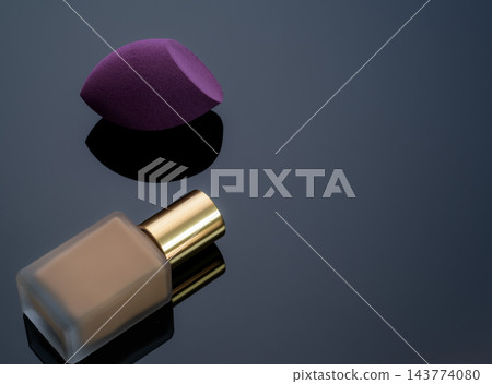 Purple makeup sponge with beige liquid foundation bottle on a dark reflective surface, studio beauty product still life for cosmetic application, makeup routine, skincare branding and advertising. Purple makeup sponge with beige liquid foundation bottle on a dark reflective surface, studio beauty product still life for cosmetic application, makeup routine, skincare branding and advertising. 143774080