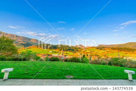 Green grass field on hill. Landscape of village in valley. Pine forest on the mountain with blue sky and white clouds on sunny day in Europe. Empty seat in the garden at the park. Small city in valley 143774539