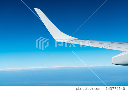 Airplane wing above a distant band of white clouds during an international daytime flight, curved white wingtip against a blue sky, a scenic passenger window view of commercial aviation and travel Airplane wing above a distant band of white clouds during an international daytime flight, curved white wingtip against a blue sky, a scenic passenger window view of commercial aviation and travel 143774540