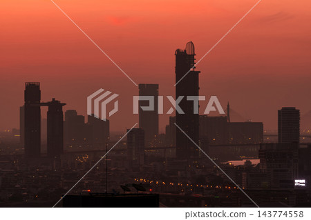 Urban skyline with high rise buildings silhouetted against a deep orange dusk sky, showing hazy metropolitan atmosphere, city lights and dense architecture for travel, pollution and evening themes 143774558