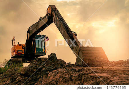 Tracked excavator parked on uneven soil at a construction site at sunset, with an extended hydraulic arm and a dirt covered bucket resting on the ground after digging, heavy equipment for earthmoving. 143774565