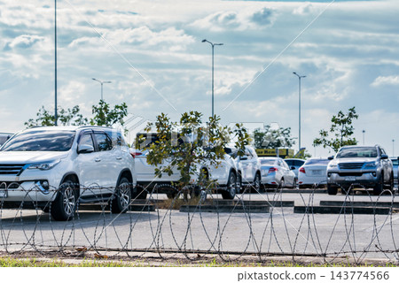 Rental cars parked behind a barbed wire fence at an airport parking lot, with white SUVs and silver sedans beside concrete wheel stops and trees, vehicle storage for an automotive rental business. Rental cars parked behind a barbed wire fence at an airport parking lot, with white SUVs and silver sedans beside concrete wheel stops and trees, vehicle storage for an automotive rental business. 143774566