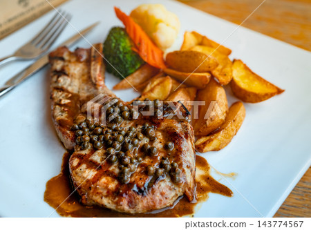 Grilled tomahawk steak with peppercorn sauce beside golden fried potato wedges and steamed vegetables, including carrot, cauliflower and broccoli, served on a white plate resting on a wooden table. 143774567