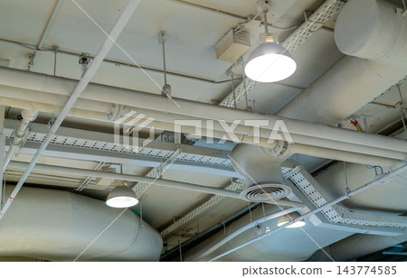 Air ducts, wiring and plumbing beneath a shopping mall ceiling, hanging lamps beside a circular ventilation diffuser, white pipes and perforated cable trays supported by rods and brackets. 143774585