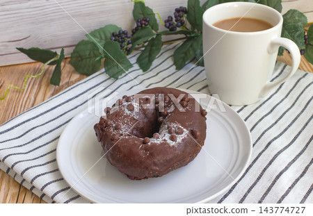 白色盤子上放著一個巧克力貝果,馬克杯裡裝著一杯拿鐵。 白色盤子上放著一個巧克力貝果,馬克杯裡裝著一杯拿鐵。 143774727