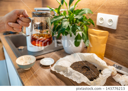 A glass teapot of amber puer tea is held above a kitchen counter. The finished citrus infused brew is separated from the leaves for serving. 143776213