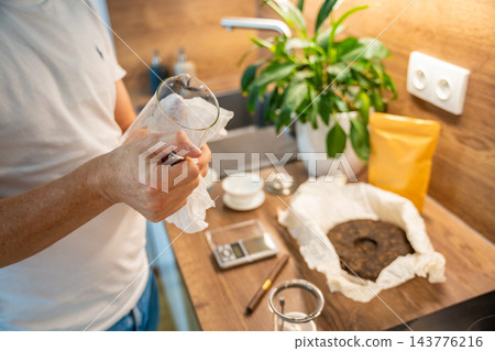 A man dries a glass tea infuser with a paper towel at a kitchen counter. Cleaning the brewing chamber prepares it for loose leaf puer tea. A man dries a glass tea infuser with a paper towel at a kitchen counter. Cleaning the brewing chamber prepares it for loose leaf puer tea. 143776216