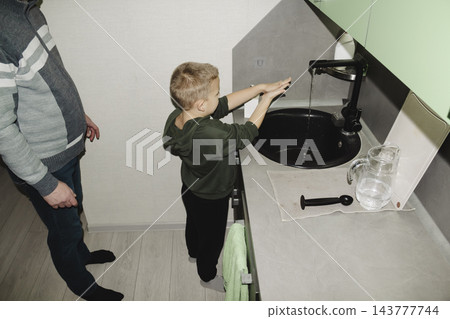 A child returns from school and, together with their dad, washes their hands and face in the sink in the modern kitchen of their home. 143777744