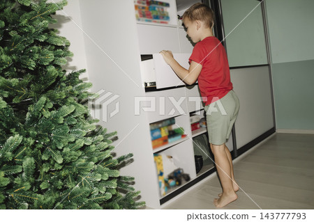 In the evening, in their cozy room, a tired child opens a white wardrobe and takes out some pajamas to change into for bed meanwhile, a Christmas tree stands in the room. In the evening, in their cozy room, a tired child opens a white wardrobe and takes out some pajamas to change into for bed meanwhile, a Christmas tree stands in the room. 143777793