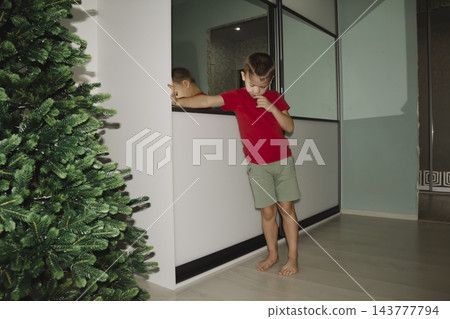 In the evening, in their cozy room, a tired child opens a white wardrobe and takes out some pajamas to change into for bed meanwhile, a Christmas tree stands in the room. In the evening, in their cozy room, a tired child opens a white wardrobe and takes out some pajamas to change into for bed meanwhile, a Christmas tree stands in the room. 143777794