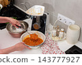 A person adds squash to a kitchen grinder while preparing squash caviar at a home kitchen counter, with a bowl, cookware, and seasonings nearby 143777927