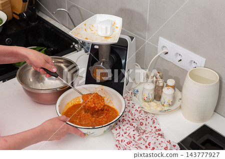 A person adds squash to a kitchen grinder while preparing squash caviar at a home kitchen counter, with a bowl, cookware, and seasonings nearby 143777927
