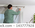 A man is hanging a curtain on the rod after washing it. It is spring, and he is doing household chores in a room with a window and some greenery 143777928