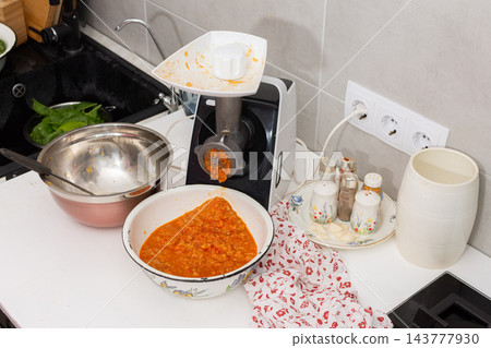A person adds squash to a kitchen grinder while preparing squash caviar at a home kitchen counter, with a bowl, cookware, and seasonings nearby 143777930