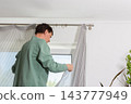 A man is hanging a curtain on the rod after washing it. It is spring, and he is doing household chores in a room with a window and some greenery 143777949