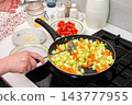 Hands stir chopped squash, carrots, and tomatoes in a pan on a kitchen stove during preparation of squash caviar at home 143777955