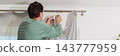 A man is hanging a curtain on the rod after washing it. It is spring, and he is doing household chores in a room with a window and some greenery, banner 143777959