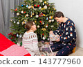 A 52-year-old couple sits on the floor, smiling and opening gifts. The room is decorated with a Christmas tree, colorful ornaments, and presents around them. It is Christmas Day 143777960