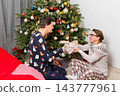A couple sits near a Christmas tree on Christmas Day. They are exchanging gifts and enjoying the holiday spirit. The room has decorations and a festive atmosphere 143777961