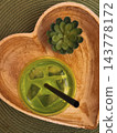 Iced green matcha tea latte with straw in heart shaped wooden tray. Top view of glass with iced matcha green tea beverage, ice cubes, straw served in heart shaped wooden tray next to small succulent 143778172