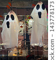 Halloween ghost decorations formed from white translucent fabric and illuminated hanging lamps feature black facial details. Ghost figures, seasonal atmosphere, party decor, event styling, background 143778173