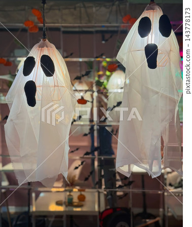 Halloween ghost decorations formed from white translucent fabric and illuminated hanging lamps feature black facial details. Ghost figures, seasonal atmosphere, party decor, event styling, background Halloween ghost decorations formed from white translucent fabric and illuminated hanging lamps feature black facial details. Ghost figures, seasonal atmosphere, party decor, event styling, background 143778173