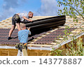 Roof installation and repair at heights with two workers fitting asphalt roofing panels over exposed framing on sloping wooden roof. 143778887