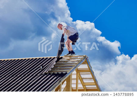 Installing corrugated asphalt roofing sheets, construction worker positions new panel beside completed section of pitched timber roof. 143778895