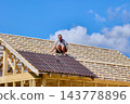 Installation of corrugated asphalt roofing sheets on timber rafters, construction worker positions panels across sloping roof during house building. 143778896