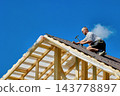 High-altitude work on timber roof with roofer using hammer to secure corrugated bitumen sheets above exposed wooden rafters. 143778897