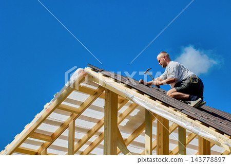 High-altitude work on timber roof with roofer using hammer to secure corrugated bitumen sheets above exposed wooden rafters. 143778897