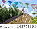 Sloped roof work involving corrugated bitumen sheet installation on timber framing with roofer holding hammer near roof ridge. 143778898