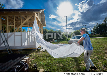 Construction wrap unrolled by builder into long sheet for covering timber frame walls with protective layer against wind and moisture. Construction wrap unrolled by builder into long sheet for covering timber frame walls with protective layer against wind and moisture. 143778899