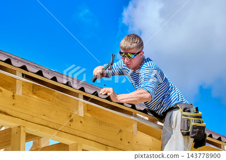 Fixing corrugated asphalt roofing sheets with hammer and nails, construction worker secures roofing along timber eaves. Fixing corrugated asphalt roofing sheets with hammer and nails, construction worker secures roofing along timber eaves. 143778900